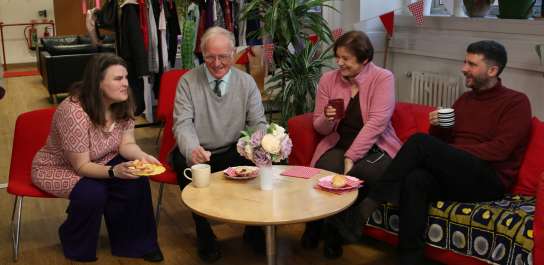 Four people sit on red chairs and sofas around a coffee table, smiling and chatting over tea and cakes in a cosy indoor setting decorated with bunting and clothing racks in the background.