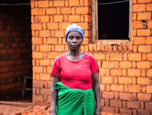 Aline Nigobora stands in front of her home, She wears a pink top and green skirt and is looking in the camera with a neutral expression