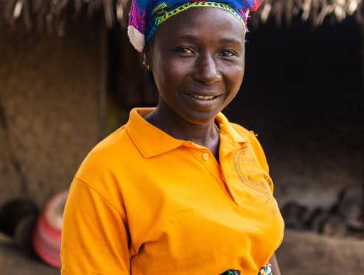 Maryama, a woman in an orange shirt and blue headscarf stands outside a thatched building her home in Sierra Leone, smiling slightly.